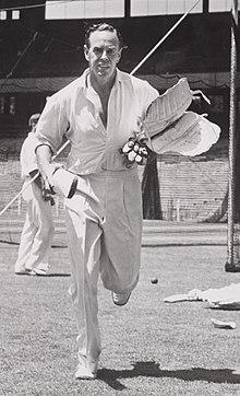 Johnson, bare-headed and wearing cricket whites, running towards the camera carrying cricket pads and gloves in his left hand and a cricket bat in his right hand