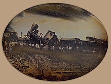 A daguerreotype of a train collision. Two trains and their locomotives can be seen in a mangled mess. A crowd of spectators can be seen observing the aftermath of the accident.