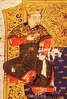 A man sits on a couch in front of a golden screen wearing brown and red robes and a gold crown.