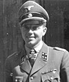 A smiling man wearing a military uniform, peaked cap and a neck order in the shape of a cross. His cap has an emblem in shape of a human skull and crossed bones.