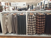 A rack of jogger pants in a shopping mall store. The sign above them reads "Drape Jogger Pants: $199. Flattering drape in casual look". Next to them is a row of plaid pajama pants.