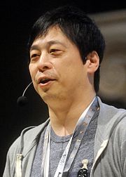 A black-haired Japanese man in his fifties at a convension, wearing a light grey t-shirt and paler grey hoodie.