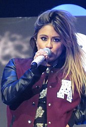 Brooke, in a brown jacket, sings into a microphone.