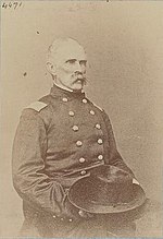 Sepia toned photo shows a seated man with a bushy moustache. He wears a dark military uniform and holds his hat in his lap.