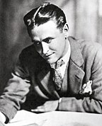 A photograph of F. Scott Fitzgerald by Nickolas Muray. Fitzgerald is bent over a desk and is examining a sheaf of papers. He is wearing a light suit and a polka-dot tie. A white handkerchief is in his breast pocket.