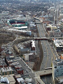 Aerial view of a divided highway in a dense urban neighborhood, passing under several bridges and near multi-story buildings and a baseball stadium