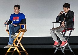 Photograph of two men seated in film director's chairs in front of a projection screen