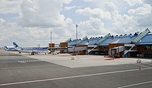 Farbfotografie in der Obersicht von einem Flugplatz. Links stehen zwei blau-weiße Flugzeuge vor dem Abfertigungsgebäude, das sich von der Bildmitte bis zum rechten Rand erstreckt.