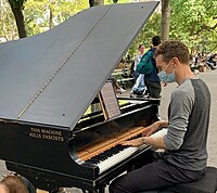 A middle-aged man plays a grand piano on a large path in a park. On the piano's side is the phrase "THIS MACHINE KILLS FASCISTS".