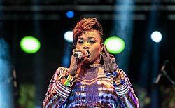Oumou Sangaré wearing a patterned outfit, singing into a microphone onstage