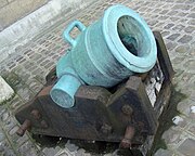 Photo of a short mortar with a wide bore. The pale green mortar rests on what looks like a rusted iron carriage.