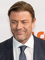 A caucasian man with short dirty blonde hair. He is looking towards the left and is wearing a suit.