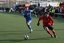 Frontale Farbfotografie von zwei Fußballspielern, die einem Ball in der Bildmitte hinterherjagen. Der linke Spieler trägt ein blaues Trikot und der rechte Spieler ein rotes Trikot und schwarze Torwarthandschuhe. Im Hintergrund steht ein kleines Publikum.