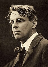 A posed black and white photograph of Yeats. He is wearing smart clothes and spectacles, while his hair looks a bit tousled