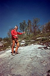A photo of Gene Cernan standing on a rock with holding a stick while participating in geology training.
