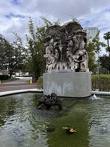 The photo depicts a fountain with a tall statue at the rear center. At the top is an eagle carving with its wings spread clutching a locked metal box. The eagle's pedestal has two people and two fish carved on either side with water spewing from the fish's mouth into the pool below. A steamship is carved into the left side and a train is carved into the right side..