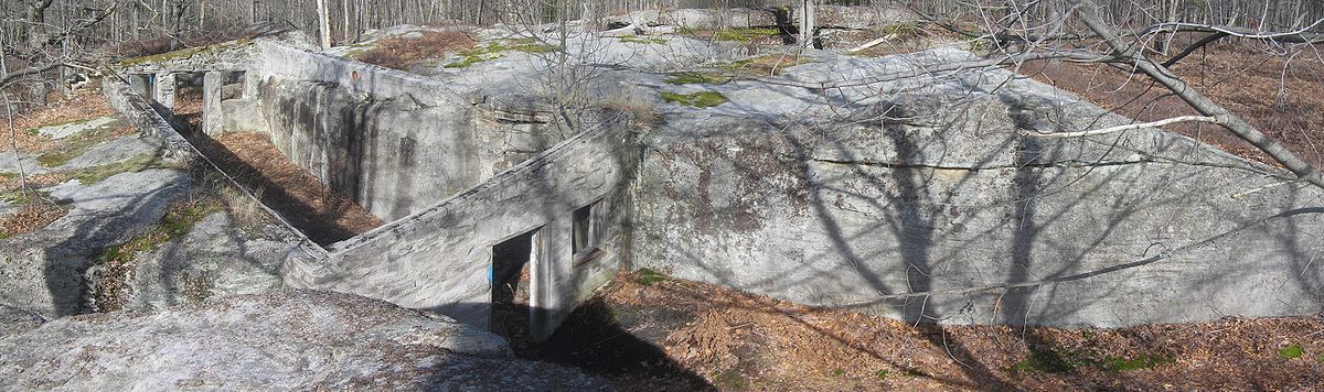 Panoramic view from above of two large boulders with walls sealing off the space between them. The walls have holes for doors and windows in them, but there is no roof or doors or glass. The ruins are at left and the far boulder runs the width of the image.
