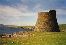 Green fields lie to the right of a body of dark blue water. At the water's edge, there is a tall, round, stone tower, possibly unroofed.