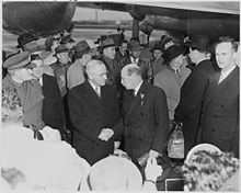 Truman shaking hands with Attlee. A large crowd surrounds them. There is a large propeller-driven airplane in the background.