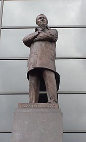 A bronze statue of a man wearing a coat with his arms folded.