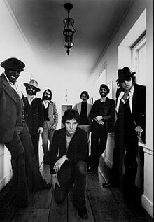 A black and white photograph of seven men standing in a hallway. One is kneeling in the center while three stand on his left and three on his right