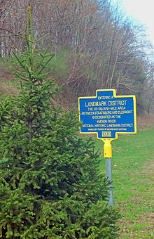 A sign with the headline "Landmark District" in yellow on a blue background, with yellow trim, behind an evergreen tree on its left. There are some trees in back.