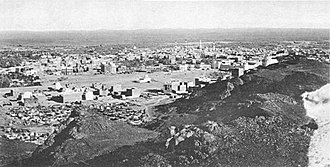 Black-and-white photograph of a city in the desert showing a basaltic ridge on the right and a skyline with numerous buildings among which is a domed mosque with two minarets
