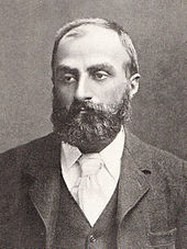 Heavily bearded man with dark receding hair, wearing a dark coloured jacket, white-collar and pale tie. He is looking slightly to the left, with a solemn expression