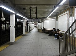 View of a wide section of the northbound platform. There is a bench (with passengers sitting on it), a stair, and a wall to the right. On the left is a row of pillars with white ceramic tile pillars containing the number "103", as well as a yellow tactile strip on the platform edge. There are subway tracks to the far left.