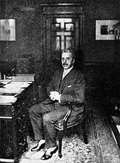 man in late middle age, dressed in a dark suit, full head of greying hair, moustached, sitting at a desk