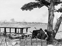 Men crouch behind the shield of an artillery gun beneath a tree overlooking a body of water