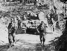 Soldiers in combat equipment carrying rifles advance along a dirt road in front of a tank