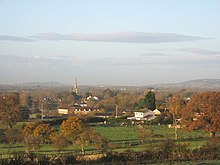 Roofs of houses showing amongst tree with prominent church tower. In the foreground are green fields with hills behind.