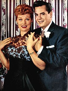 Colored glamorous shot of Lucille Ball and Arnaz standing: Both are smiling to the front. Ball at the left wears a ceremonial gown; Arnaz at right wears a tuxedo.