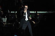 A man wearing a black suit, pants, and sunglasses is holding a microphone on his right hand