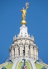 A stone, columned cupola atop a green, tiled-dome. The cupola is topped with a gold globe, upon which stands a gold statue with an outstretched arm and holding a staff.