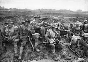 Black and white photo of six men wearing military uniform seated on a muddy slope in France, December 1916. Unidentified members of the Australian 5th Division, enjoying a "smoko" near Mametz, on the Somme. Some are wearing slouch hats, steel helmets, sheepskin jackets and woollen gloves, demonstrating both the variety of official battledress, and how it was modified and augmented, for local conditions.