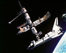 A cluster of cylindrical modules with projecting feathery solar arrays and a spaceplane docked to the lower module. In the background is the blackness of space, and in the lower right corner is Earth.