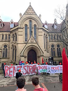 Protesters standing in front of the John Owens Building on the University of Manchester campus holding banners that read "RENT STRIKE" and "CROSS A PICKET? WHAT A TORY!". Another protester is speaking through a megaphone.