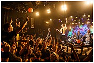 Farbfotografie von einem Konzert auf einer kleinen Bühne. Links jubelt das Publikum mit erhobenen Armen und rechts spielen vier Musiker, bestehend aus zwei Gitarristen, einem Schlagzeuger und einem Sänger. Der Sänger hält den Mikrofonständer ins Publikum. Im Hintergrund sind ein Graffiti-Bild, die Lautsprecher und Scheinwerfer.