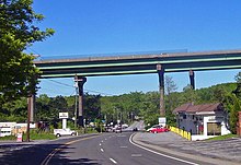 A pair of bridges pass from left to right on tall supports that tower over the other structures in the area, such as telephone poles and single-story buildings. The two-lane NY 22 passes underneath the bridge and proceeds into the background.