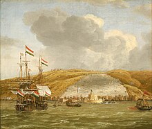 Two ships with sails and some smaller boats with oars in a harbor, with a walled city and a citadel behind them and a steep hill in the background