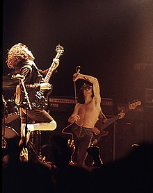 Angus, about 24, in mid-leap, right leg cocked, back arched while playing guitar. Scott holds microphone above his head, another tattoo partly visible.
