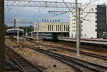 Frontale Farbfotografie von einem Bahnhof mit Gleisen im kompletten Vordergrund und Bahnsteige im hinteren linken Bereich und rechts. Im Hintergrund sind eine überdachte Halle und zwei große Gebäude, von denen auf dem linken Dach Tallinn steht. In der Bildmitte steht ein älterer Zug.
