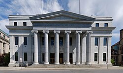 A three-story light-colored stone building. In the front a pedimented central pavilion with six Ionic columns projects. Between the second and third stories of the main facade there is a large molded cornice.