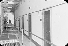 "Black and white photograph of a landing in Kilmainham Jail, with closed cell doors visible on either side and two uniformed guards standing towards the left"