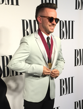 Ricky Reed in a white suit and sunglasses. The text "BMI" in written several times on the wall behind him.