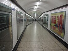 A long corridor with tiled floor and walls and a curved ceiling. The walls are lined at regular intervals with large posters framed behind glass