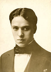 Frontales Schwarzweißporträt eines jungen Mannes mit ernstem Blick. Er trägt wellige Haare mit Mittelscheitel und einen dunklen Anzug mit Fliege.