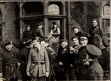 "Black and white photograph of a group of approximately sixteen men in military police uniform outside a house with a broken window through which a civilian can be seen"